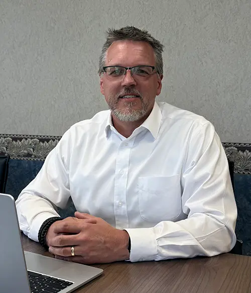 Headshot of Brant Darnell, wearing a white dress-shirt, sitting at conference table in front of a laptop.