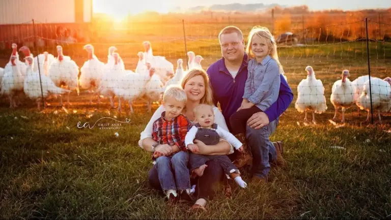 Family of five sitting on a grassy area in front of chicken-wire fencing. Behind the fencing is a flock of turkeys and behind all of them is a field with the sun setting in the background.