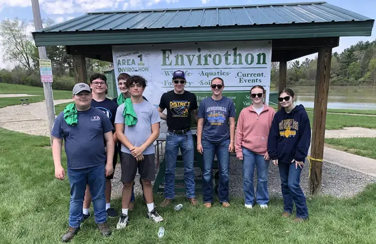 Eight Ayersville high school students stand grouped in front of a small park shelter, displaying a sign for the Envirothon. Envirothon Photo from left to right: Hayden Gorrell, Zachary Stiltner, Levi Miller, Kaysen Hattemer, Levi Ankney, Adilae Comden, Ava Sandleben, Olivia Hattemer.
