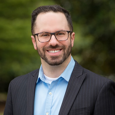 Professional headshot of a smiling man with short dark hair, glasses, and a beard, wearing a pinstriped suit jacket over a light blue shirt, photographed outdoors against a blurred green background.