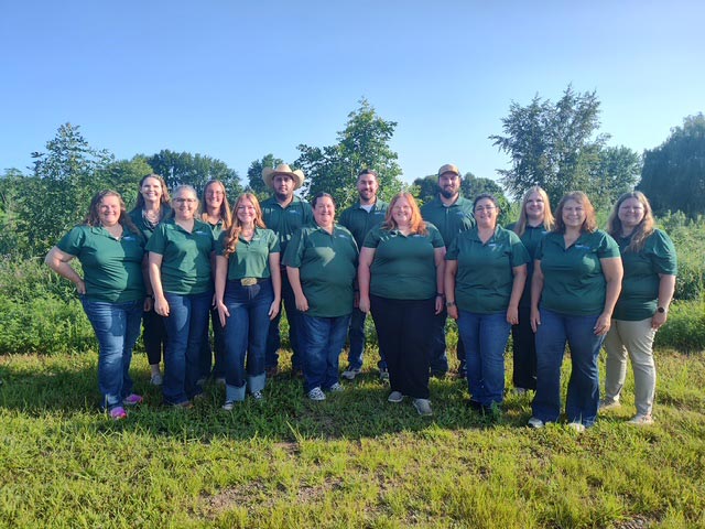 A group of about 14 people wearing matching dark green polo shirts and jeans pose together outdoors in a grassy field with trees under a clear blue sky.