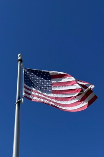 An American flag flying on a metal flagpole, waving against a clear deep blue sky.