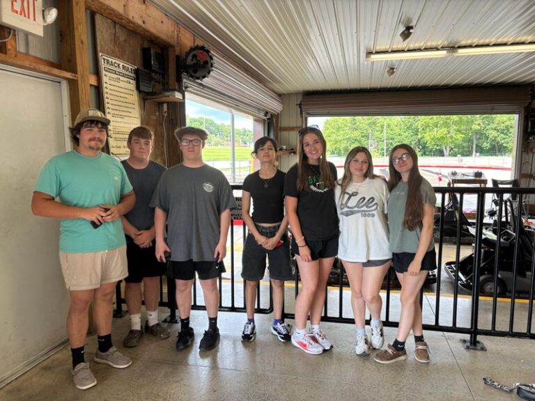 A group of seven teenagers pose together inside a go-kart track garage, with track rules posted on the wall and go-karts visible in the background.