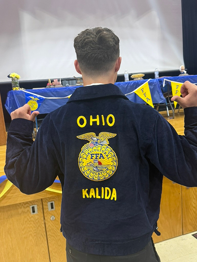 Back view of a young man wearing a navy blue corduroy FFA jacket embroidered with "Ohio" above the National FFA Organization emblem and "Kalida" below it, standing in a gymnasium decorated with FFA pennants near a table set up for an event.