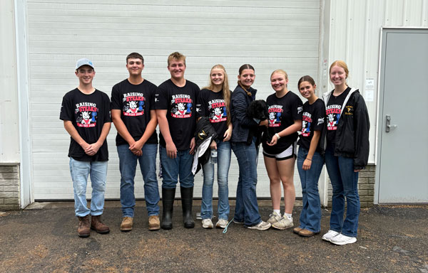 A group of eight teenagers wearing matching black "Raising the Steaks: Ag Fest 2026" t-shirts pose together in front of a rabbit barn, with one girl holding a small black dog.