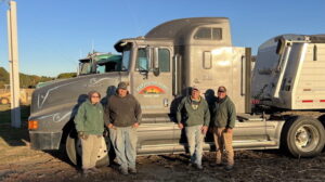 Four people stand together in front of a gray semi-truck bearing the "Maranatha Acres, Delta, Ohio" logo, parked outdoors near a grain trailer under a clear blue sky.