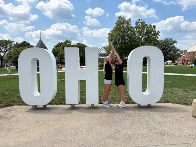 Two young women pose playfully between large white letters spelling "OHIO" in an outdoor park setting under a partly cloudy sky.