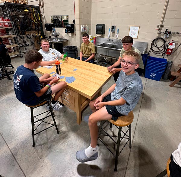 A group of teenage boys sit on stools around a wooden workbench in a shop classroom, playing cards together while one smiles at the camera in the foreground.