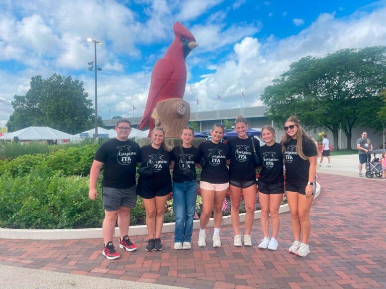 A group of seven people wearing matching black "Evergreen FFA" t-shirts pose together in front of a large red cardinal statue at an outdoor fairground plaza.