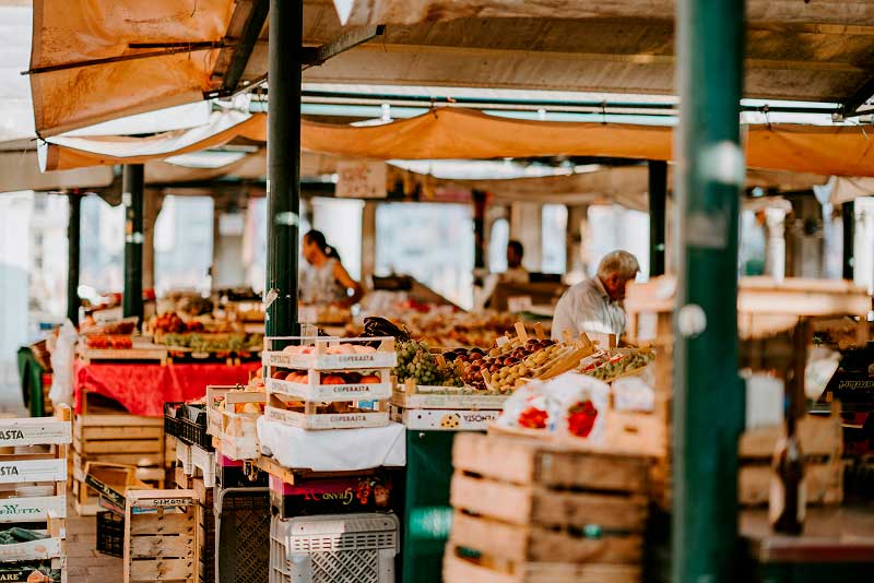 An outdoor covered market with wooden crates of fresh produce, including grapes and vegetables, as vendors and shoppers move between the stalls under canvas awnings.