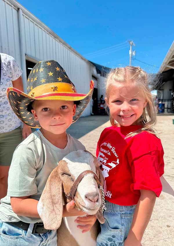 A young boy wearing a star-patterned cowboy hat holds a tan and white goat by its lead, standing next to a smiling young girl in a red "Wee Wee Show Ottawa Co." t-shirt, at an outdoor livestock show.