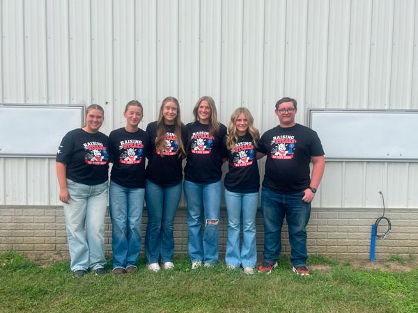 Six teens, five girls and one boy, stand shoulder to shoulder in a row on grass in front of a white metal building. They all wear matching black T-shirts with a colorful cartoon graphic and the words "Raising Steaks." Most of them wear jeans, and they are smiling.