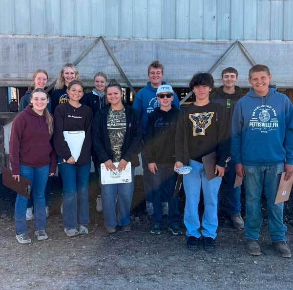 Eleven students in sweatshirts and hoodies stand in two rows outside a metal farm building, several holding clipboards.