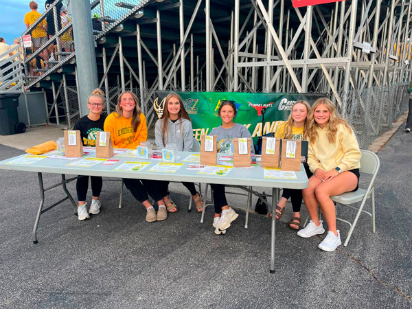 Five teenage girls sit and stand behind a folding table at a football stadium, with bleachers and a green sponsor banner behind them. The table holds brown paper bags with labels and colorful printed flyers. The girls wear casual school-spirit clothes in green, gold, and gray, and most are smiling at the camera.