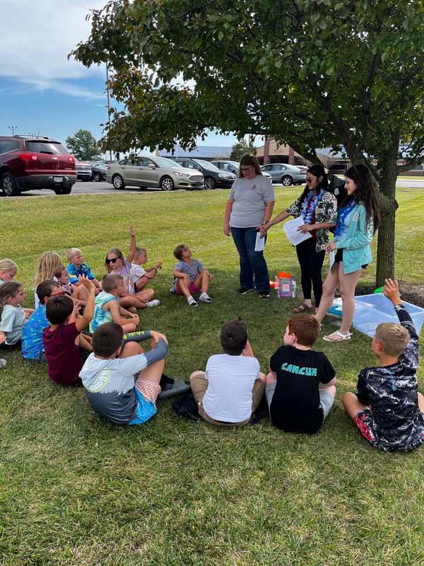 A group of about 15 children sit in a circle on a lawn under a tree, and several raise their hands. Three adults stand together at the edge of the circle: a woman in a gray shirt and jeans, and two younger women holding papers and wearing colorful leis. A clear plastic tub and a small container sit on the grass nearby, and parked cars and a brick building are visible in the background.