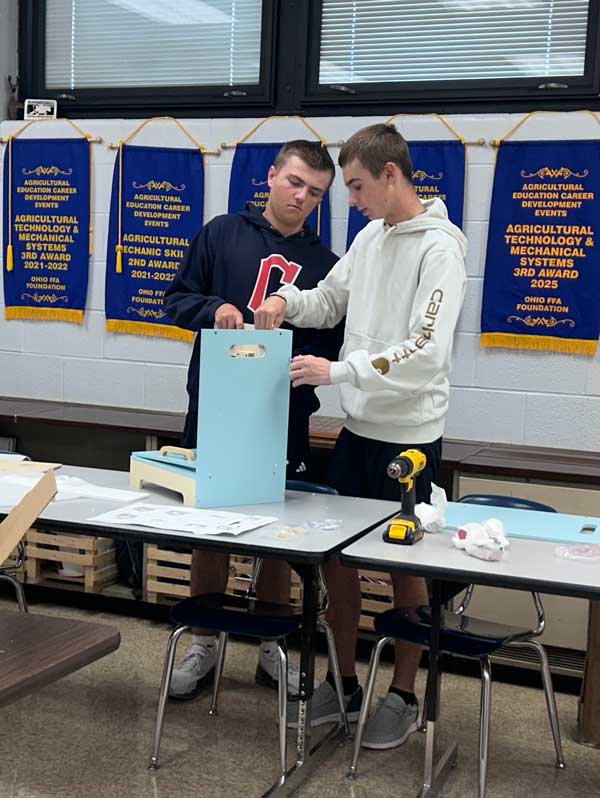 Two teenage boys stand at school tables and assemble a light blue wooden project. One boy in a navy Cleveland “C” sweatshirt holds the blue panel upright while the other, in a white Carhartt hoodie, steadies it. A yellow cordless drill and printed instructions sit on the table. Blue Ohio FFA award banners for agricultural technology and mechanical systems hang on the wall behind them.