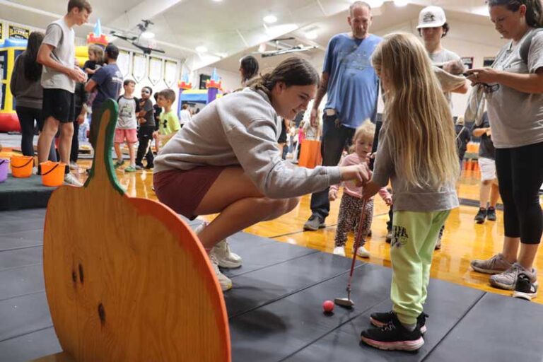 A young woman in a gray sweatshirt crouches on a black mat in a school gymnasium and helps a little girl with long blonde hair hold a toy putter. A red ball sits in front of the club, and a wooden mini-golf obstacle with a green top fills the left foreground. Behind them, a toddler stands nearby, adults and children mill around the gym floor, and inflatable bounce houses line the back wall.