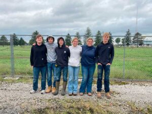 Six teens stand in a row in front of a chain-link fence, with evergreen trees and a cloudy sky behind them.