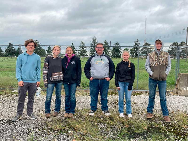 Six teens stand arm in arm in front of a chain-link fence on a gravel lot under a cloudy sky.