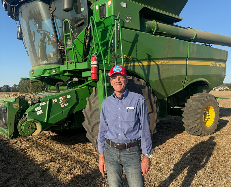 A smiling man in a red baseball cap, blue button-up shirt, and jeans stands in a harvested field in front of a large green combine. The combine’s cab, ladder, and large tire with a yellow wheel are visible behind him, and the sky is clear and blue.