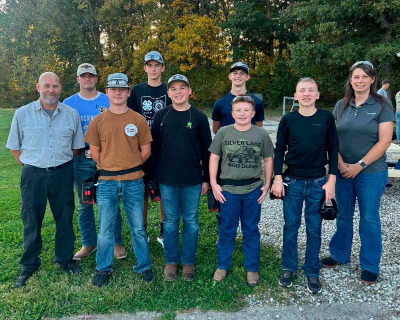 Nine people, seven teens and two adults, pose on grass and gravel in front of fall trees, several wearing ball caps.